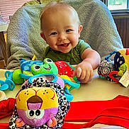 Candor is registered to the contest to win money with this photo: baby, smiling, high_chair, toy, plush_toy, indoor, child, happy, face, person, green_clothing, table, window, curtain, furniture, hand, head, colorful, seat, infant