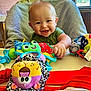 baby, smiling, high_chair, toy, plush_toy, indoor, child, happy, face, person, green_clothing, table, window, curtain, furniture, hand, head, colorful, seat, infant