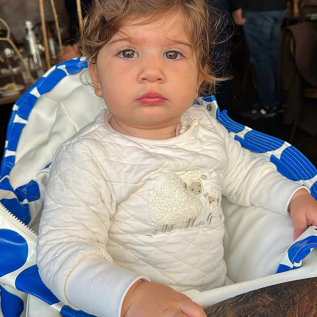 Kaïa a rejoint le concours — aidez-le/la à gagner de superbes lots ! baby, background_people, child, clothing, curly_hair, cute, earrings, expression, face, furniture, hand, high_chair, indoor, person, portrait, seated, table, toddler, white_sweater, wood
