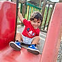 Timothée a rejoint le concours — aidez-le/la à gagner de superbes lots ! blue_shoes, child, curly_hair, daylight, fun, grass, green_fence, happy, outdoor, park, person, play, playground, red_shirt, shorts, slide, smiling, sneakers, toddler, wooden_structure