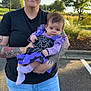 baby, bat_design, black_tshirt, casual_wear, cute_outfit, daylight, facial_expression, holding, infant, jeans, nature, outdoor, parking_lot, person, purple_clothing, sidewalk, sunglasses, tattoos, trees, woman