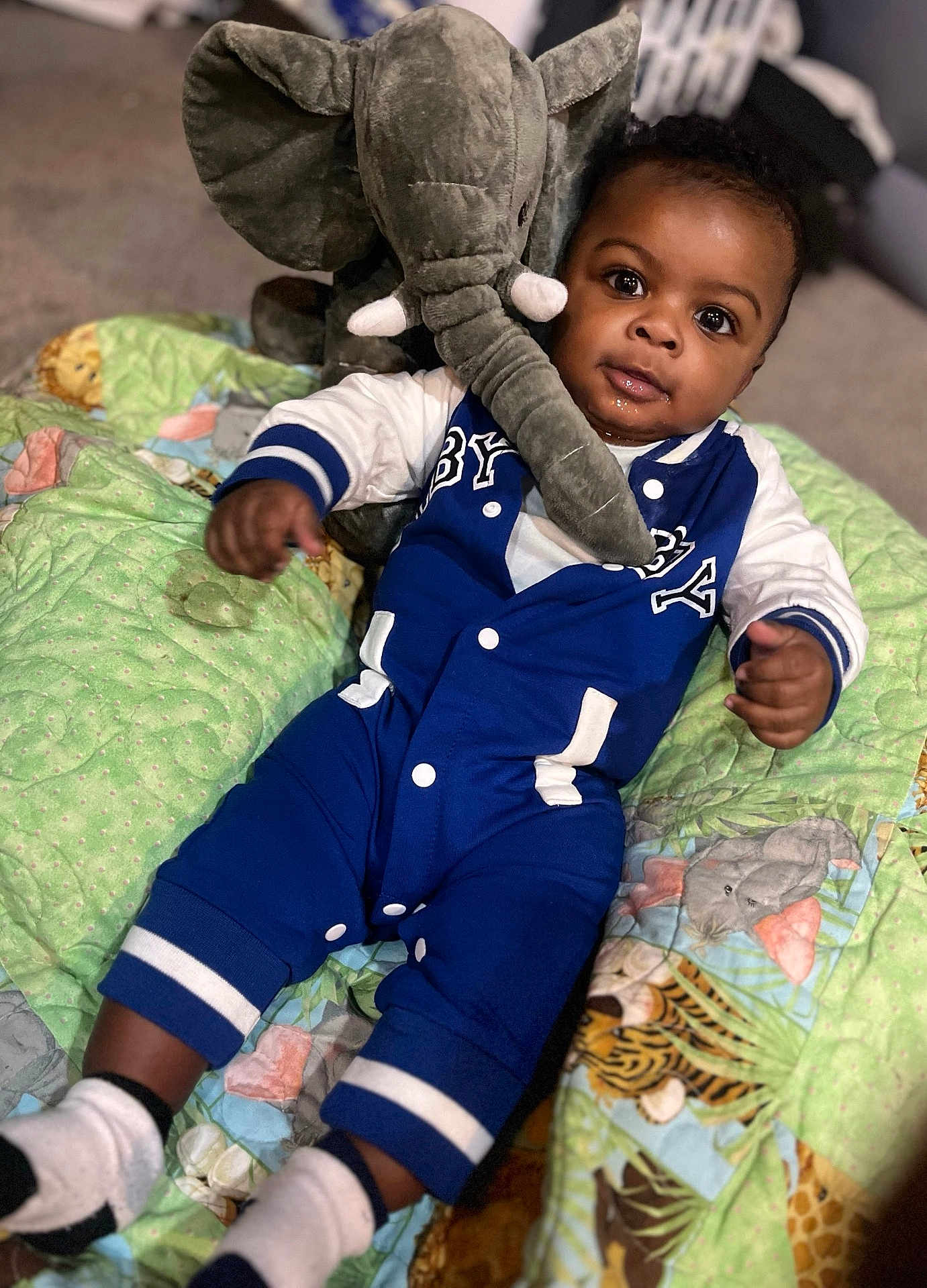 Samorion is registered to the contest to win money with this photo: animal_print, baby, blue_outfit, carpet, child, colorful, cozy, cute, drool, indoor, infant, lying_down, plush_elephant, portrait, quilt, smiling, socks, soft_toy, toy, white_sleeves