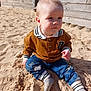 toddler, child, sand, playground, outdoor, sunny, casual_clothing, brown_sweater, blue_pants, socks, sand_dust, curious, sitting, daylight, wooden_fence, background_people, young_child, face, portrait, casual