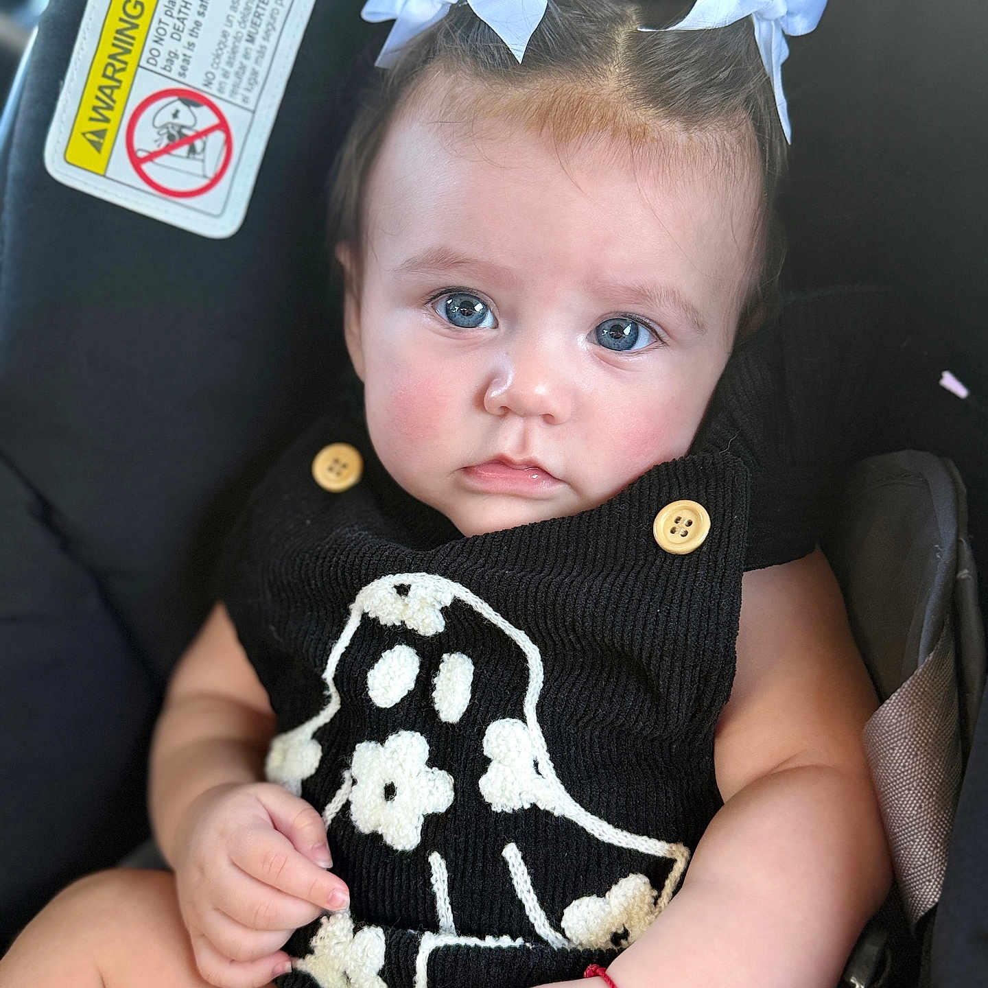 Mackenzie joined the competition — help win amazing prizes! baby, child, infant, car_seat, blue_eyes, white_bows, black_outfit, ghost_design, flower_pattern, close_up, portrait, cute, adorable, soft_lighting, hand, face, person, sitting, indoor, young