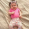 baby, infant, smiling, pink_clothing, headband, bed, blanket, cute, happy, lying_down, patterned_pants, nike_logo, child, indoors, soft_lighting, portrait, one_person, young_child, human, headwear