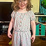 child, girl, curly_hair, romper, striped_clothing, floral_pattern, smile, indoor, wood_floor, toy_house, television, playful, person, standing, messy_hair, room, head, face, furniture, home