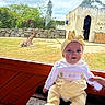 Anna a rejoint le concours — aidez-le/la à gagner de superbes lots ! baby, child, infant, yellow_clothing, headband, bow, bench, wood, window, giraffe, animal, grass, stone_wall, building, sky, clouds, outdoor, zoo, person, hand
