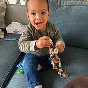 Isaac a rejoint le concours — aidez-le/la à gagner de superbes lots ! blue_couch, chain, child, clothing, colorful, couch, curly_hair, face, furniture, happy, home, indoor, jeans, person, sitting, smiling, sock, tissue_box, toddler, toy