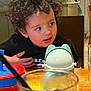 toddler, child, curly_hair, black_shirt, table, sippy_cup, frog_design, glass, straw, dining, indoor, wooden_table, curious_expression, face, person, seat, restaurant, plastic_cup, young_child, casual
