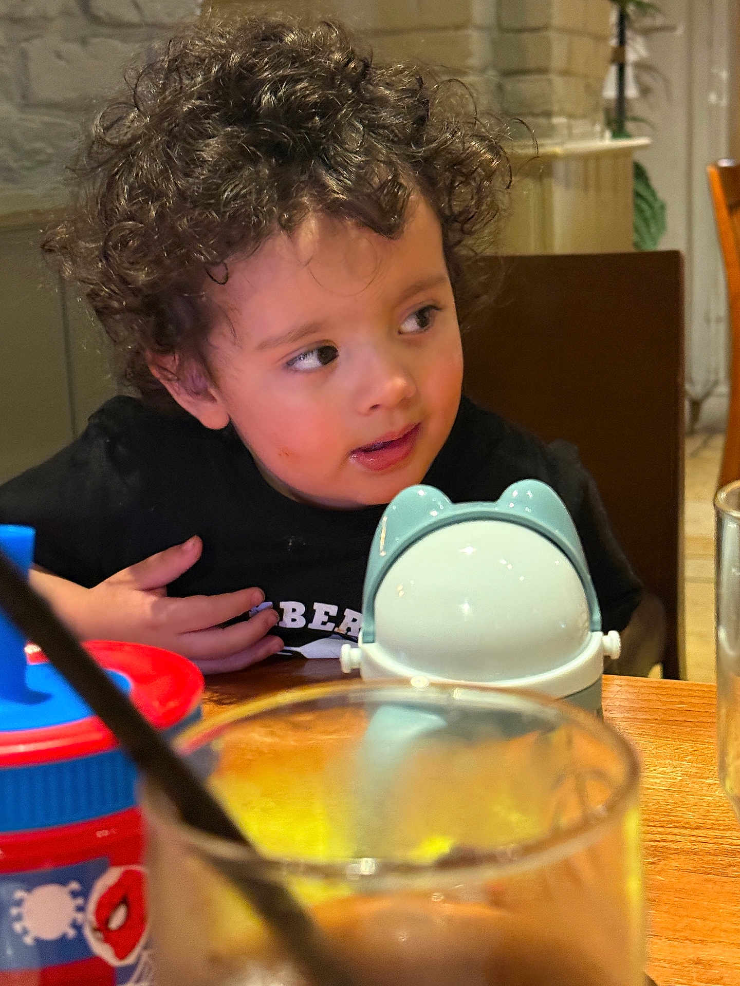Shivan is registered to the contest to win money with this photo: toddler, child, curly_hair, black_shirt, table, sippy_cup, frog_design, glass, straw, dining, indoor, wooden_table, curious_expression, face, person, seat, restaurant, plastic_cup, young_child, casual