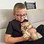 boy, child, glasses, puppy, dog, pomeranian, couch, indoor, black_clothing, light_hair, pet, cute, fluffy, holding, seated, relaxed, home, cozy, affection, smiling