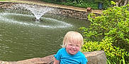 Nova is registered to the contest to win money with this photo: child, toddler, blonde_hair, blue_shirt, shorts, sneakers, stone_bench, pond, water_fountain, greenery, trees, bushes, outdoor, nature, smiling, sitting, playful, sunny, daytime, park