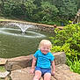 child, toddler, blonde_hair, blue_shirt, shorts, sneakers, stone_bench, pond, water_fountain, greenery, trees, bushes, outdoor, nature, smiling, sitting, playful, sunny, daytime, park