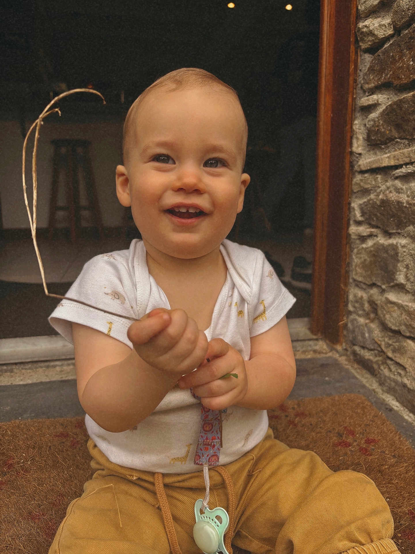 Telio a rejoint le concours — aidez-le/la à gagner de superbes lots ! toddler, child, smiling, sitting, doormat, stone_wall, doorway, pacifier, white_shirt, yellow_pants, twig, indoor, outdoor, happy, cute, baby, portrait, hand, face, eyes