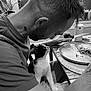 black_and_white, cat, tuxedo_cat, man, dining_table, food, plate, knife, indoor, window, plant, curious, tattoo, short_hair, seated, looking_at_each_other, intimate, home, casual_clothing, close_up