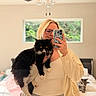 person, cat, black_and_white_cat, bedroom, mirror_selfie, glasses, blonde_hair, ceiling_fan, window, bed, gray_cat, casual_clothing, smartphone, indoor, furniture, relaxed, pet, holding, home, reflection