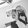 person, cat, mirror, selfie, black_and_white, glasses, smartphone, tuxedo_cat, whiskers, long_hair, ceiling_fan, window, bed, blanket, indoor, hand, sleeve, home, furniture, reflection