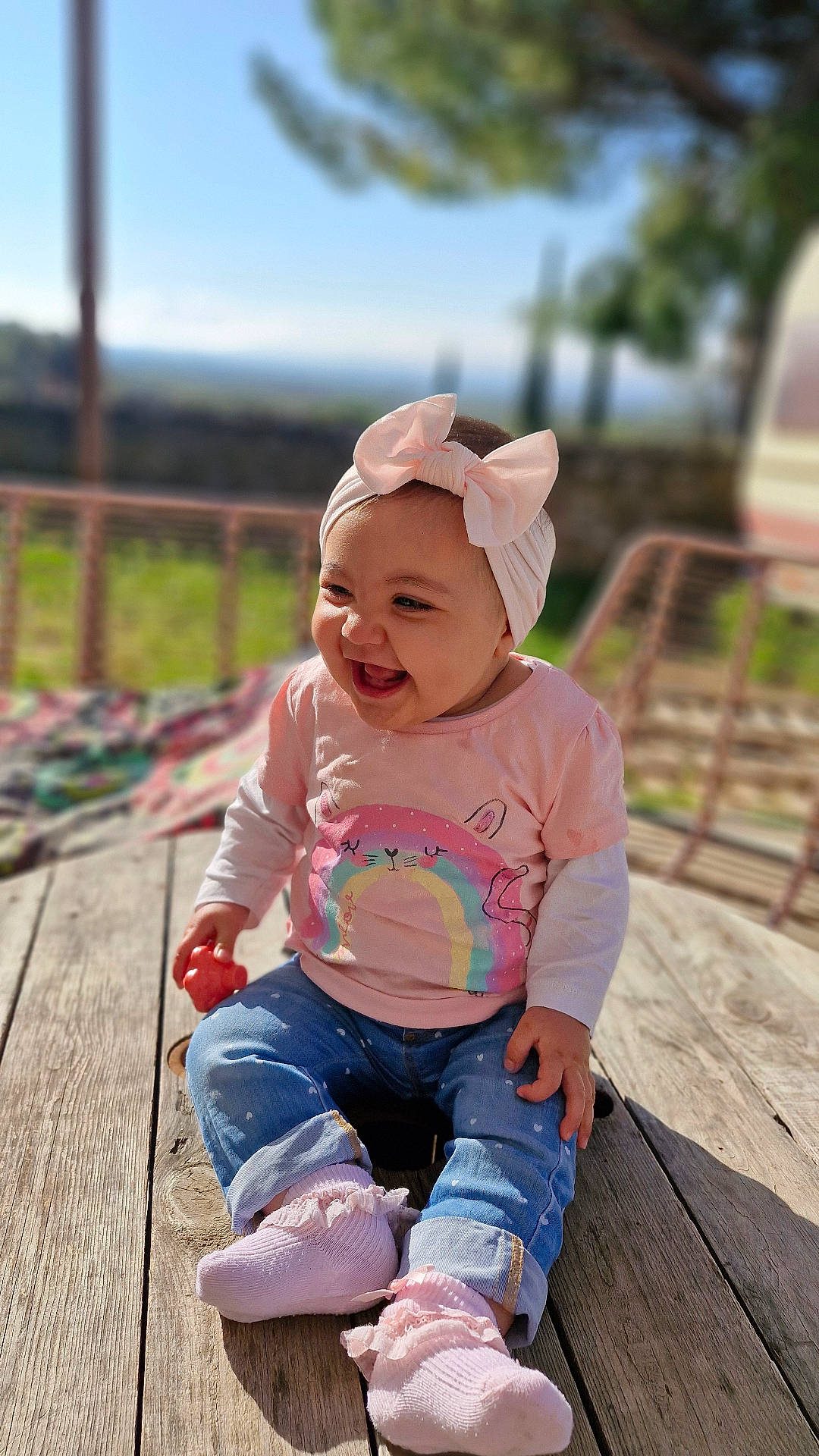 Eliana a rejoint le concours — aidez-le/la à gagner de superbes lots ! baby, bow, cat_design, child, cute, daylight, happy, headband, infant, jeans, nature, outdoor, pink, portrait, rainbow, sitting, smiling, socks, sunlight, wooden_surface