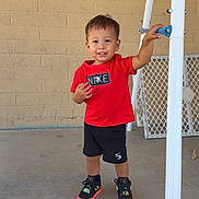 Angel joined the competition — help win amazing prizes! toddler, child, standing, red_shirt, black_shorts, sneakers, concrete_floor, brick_wall, white_pole, blue_handle, smiling, outdoor, casual_clothing, young_child, person, happy, daylight, footwear, playing, leaning