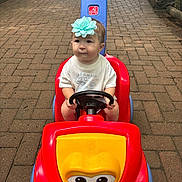 Isabelle is registered to the contest to win money with this photo: toddler, child, toy_car, headband, flower, steering_wheel, outdoor, pavement, brick_path, red, yellow, blue, person, play, cute, baby, summer, casual_clothing, vehicle, sidewalk