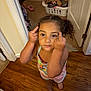 child, barefoot, wooden_floor, laundry_basket, door, carpet, indoor, person, clothing, hair, expression, hand_gesture, footwear, room, furniture, casual, curly_hair, pink, standing, flooring