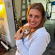 Apricot is registered to the contest to win money with this photo: animal, casual, cat, concrete_floor, door, garage, happy, holding, house, kitten, ladder, orange_cat, outdoor, pet, plant, portrait, smiling, sunset_light, white_hoodie, woman