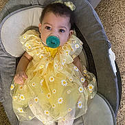 Jordynn is registered to the contest to win money with this photo: baby, child, pacifier, yellow_dress, daisy_pattern, baby_seat, carpet, infant, cute, comfort, indoors, toddler, sitting, clothing, face, headband, white_tights, looking_at_camera, soft_furniture, relaxed