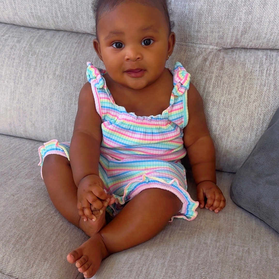 Kalia is registered to the contest to win money with this photo: baby, bright_eyes, child, clothing, comfortable, couch, curly_hair, cute, face, feet, hands, home, indoors, infant, person, seated, skin, smile, sofa, striped_outfit