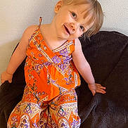 Tessa a rejoint le concours — aidez-le/la à gagner de superbes lots ! toddler, child, smile, orange_clothing, patterned_clothing, barefoot, blanket, sitting, indoor, wall, happy, cute, young_child, head_tilt, playful, person, portrait, colorful, feet, fabric