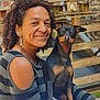 woman, dog, smile, curly_hair, earrings, outdoor, brick_wall, wooden_pallet, table, phone, pet, black_dog, tan_markings, casual_clothing, friendly, seated, daylight, portrait, happy, companionship