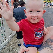 Bruce is registered to the contest to win money with this photo: adult_hand, background, casual_clothing, child, daylight, fence, happy, outdoor, park, pavement, people, person, red_shirt, shorts, smiling, summer, tattoo, toddler, trees, waving_hand