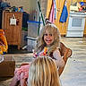 child, children, girl, toddler, smiling, happy, portrait, blonde_hair, long_hair, sitting, chair, kitchen, home, indoor, wooden_cabinets, stove, toys, apron, cardboard_box, family
