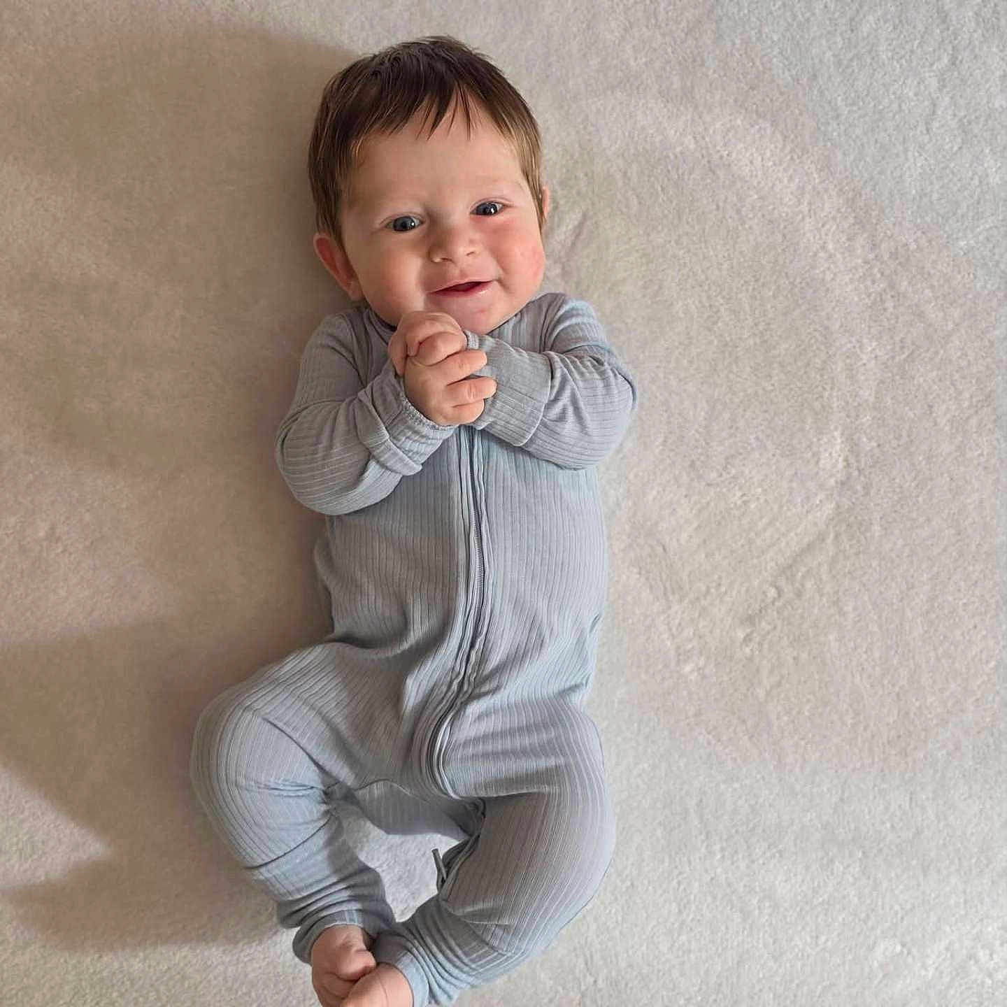 James joined the competition — help win amazing prizes! baby, infant, child, onesie, smile, hands, feet, lying_down, blue_clothing, soft_texture, face, happy, cute, portrait, human, young_child, indoors, skin, head, expression