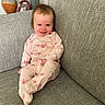 baby, infant, smiling, onesie, pink_clothing, couch, gray_furniture, living_room, portrait, happy, child, indoor, seated, cozy, decor, vase, photo_frame, home, cute, relaxed