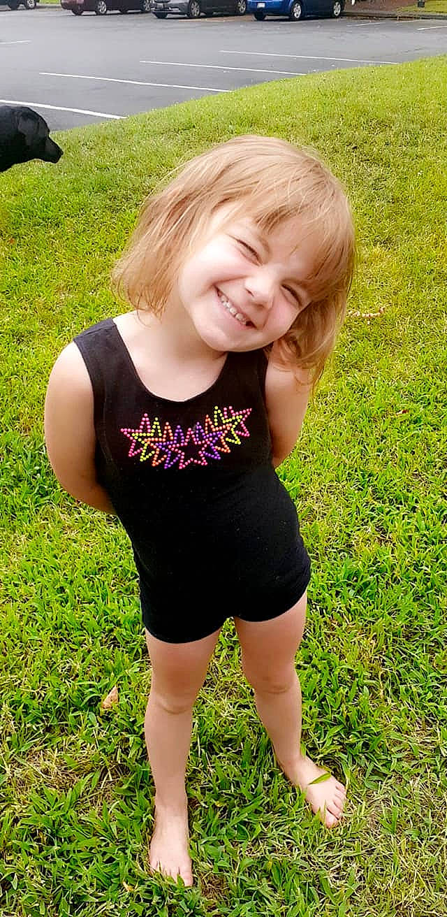 Lily is registered to the contest to win money with this photo: active_tank, baby_toddler_clothing, face, grass, hair, hairstyle, happy, head, joy, leg, people, people_in_nature, person, plant, sleeve, smile, sportswear, summer, t_shirt, thigh