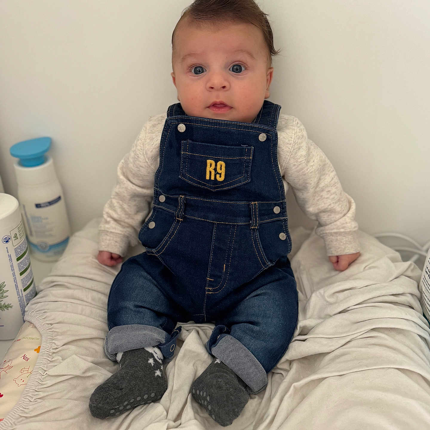 Maylonn participe au concours pour gagner de l'argent avec cette photo : baby, baby_care, blanket, blue_eyes, bottle, child, clothing, curious, cute, denim_overalls, expression, face, hair, indoor, infant, person, sitting, socks, soft_toys, white_background