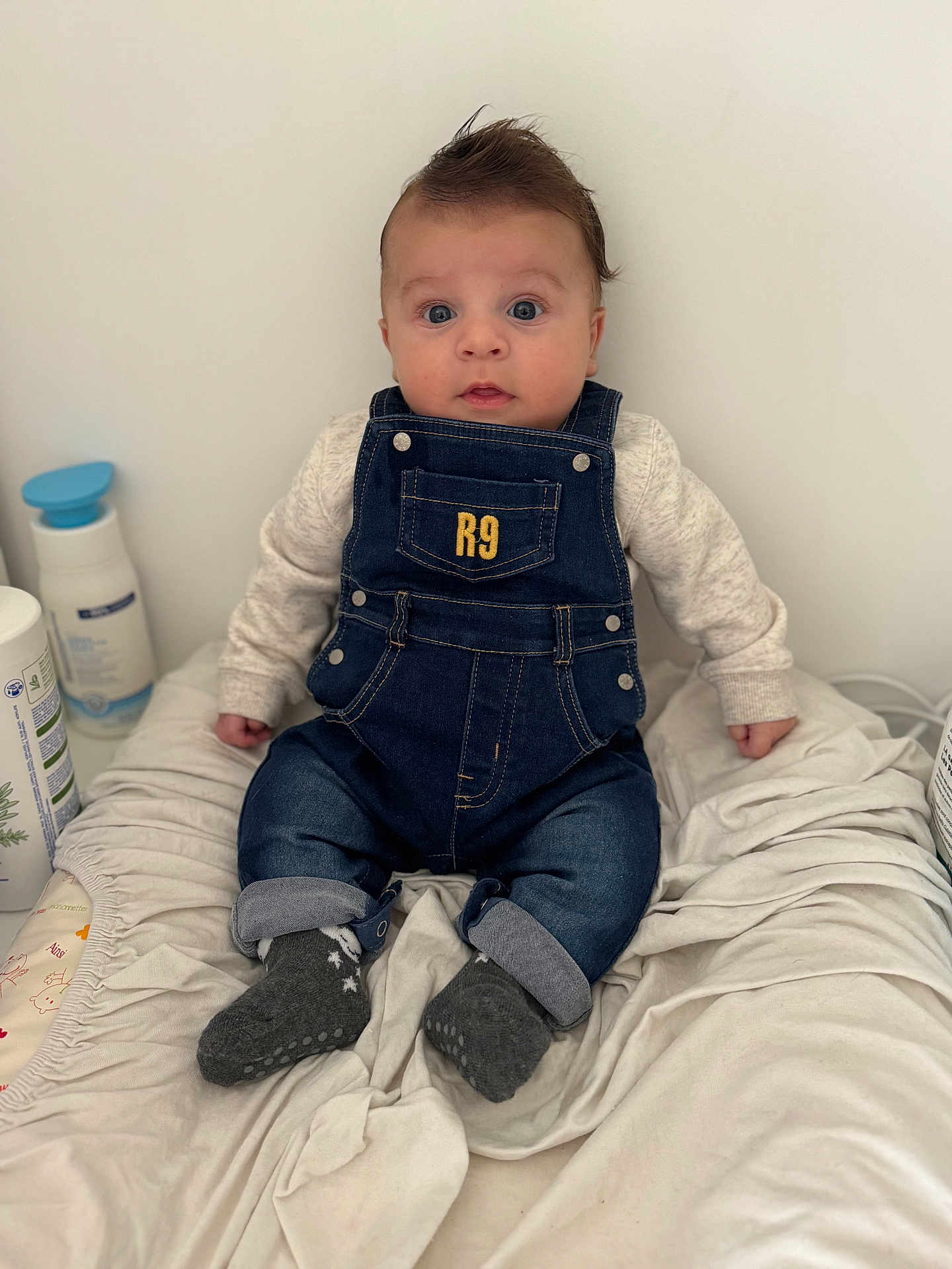Maylonn participe au concours pour gagner de l'argent avec cette photo : baby, denim_overalls, socks, blanket, infant, clothing, face, person, cute, indoor, expression, hair, child, sitting, blue_eyes, soft_toys, baby_care, bottle, white_background, curious