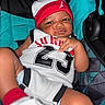 Acaio is registered to the contest to win money with this photo: baby, child, infant, car_seat, hat, jersey, number_23, clothing, red, white, black, patterned, seat, expression, face, skin, cute, young, person, indoors