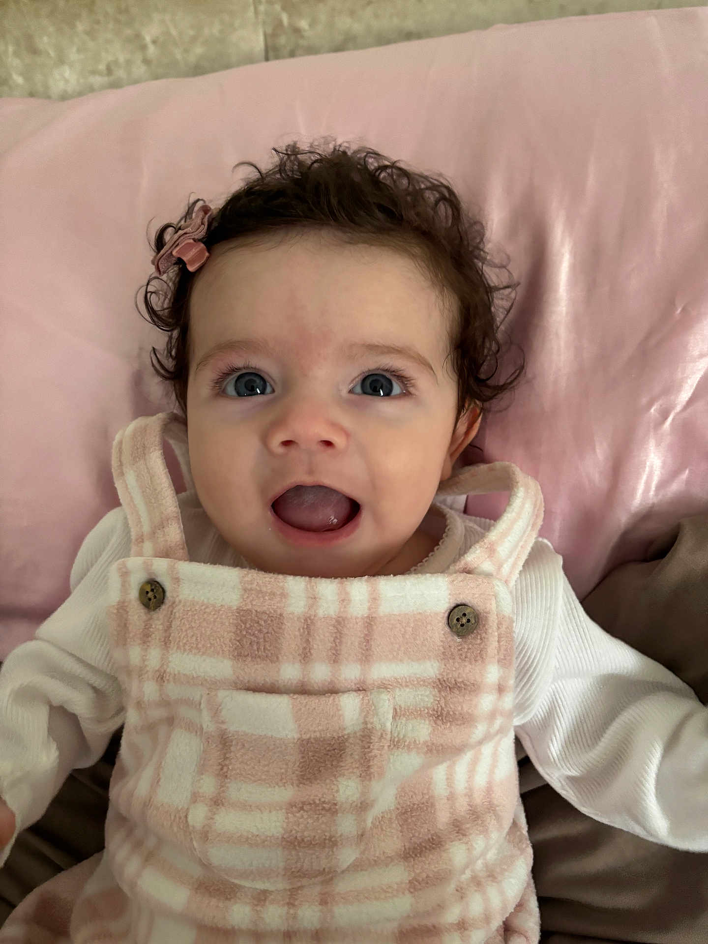 Aria is registered to the contest to win money with this photo: baby, child, infant, curly_hair, blue_eyes, pink_outfit, plaid_pattern, smiling, open_mouth, cute, lying_down, soft_texture, cushion, buttoned_clothing, happy, indoors, portrait, person, head_accessory, cozy