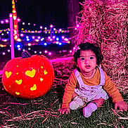 Tashvi is registered to the contest to win money with this photo: toddler, child, pumpkin, carved_pumpkin, grass, hay, night, string_lights, colorful_lights, festival, outdoor, cute, smiling, sitting, clothing, face, head, celebration, autumn, festive