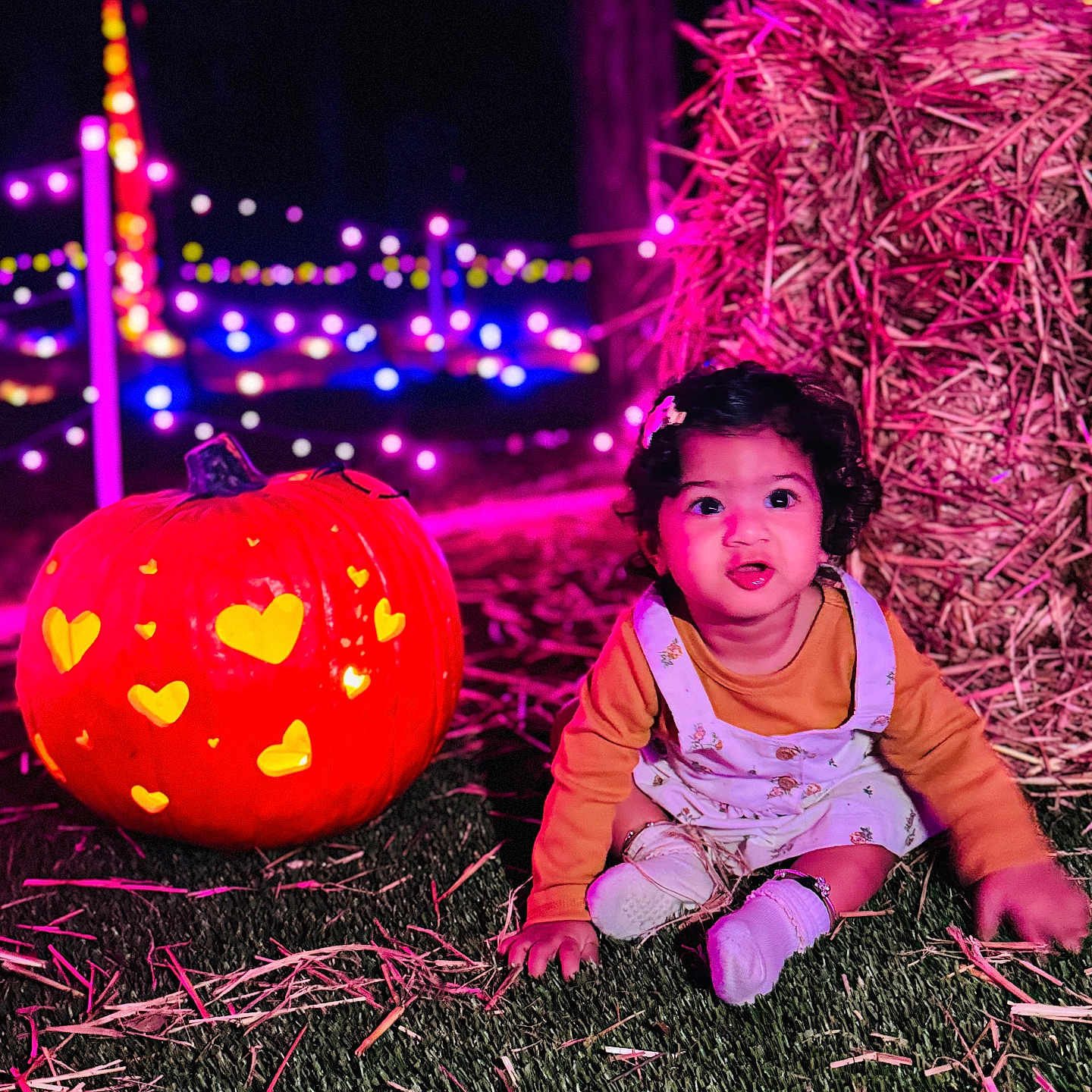 Tashvi is registered to the contest to win money with this photo: autumn, carved_pumpkin, celebration, child, clothing, colorful_lights, cute, face, festival, festive, grass, hay, head, night, outdoor, pumpkin, sitting, smiling, string_lights, toddler
