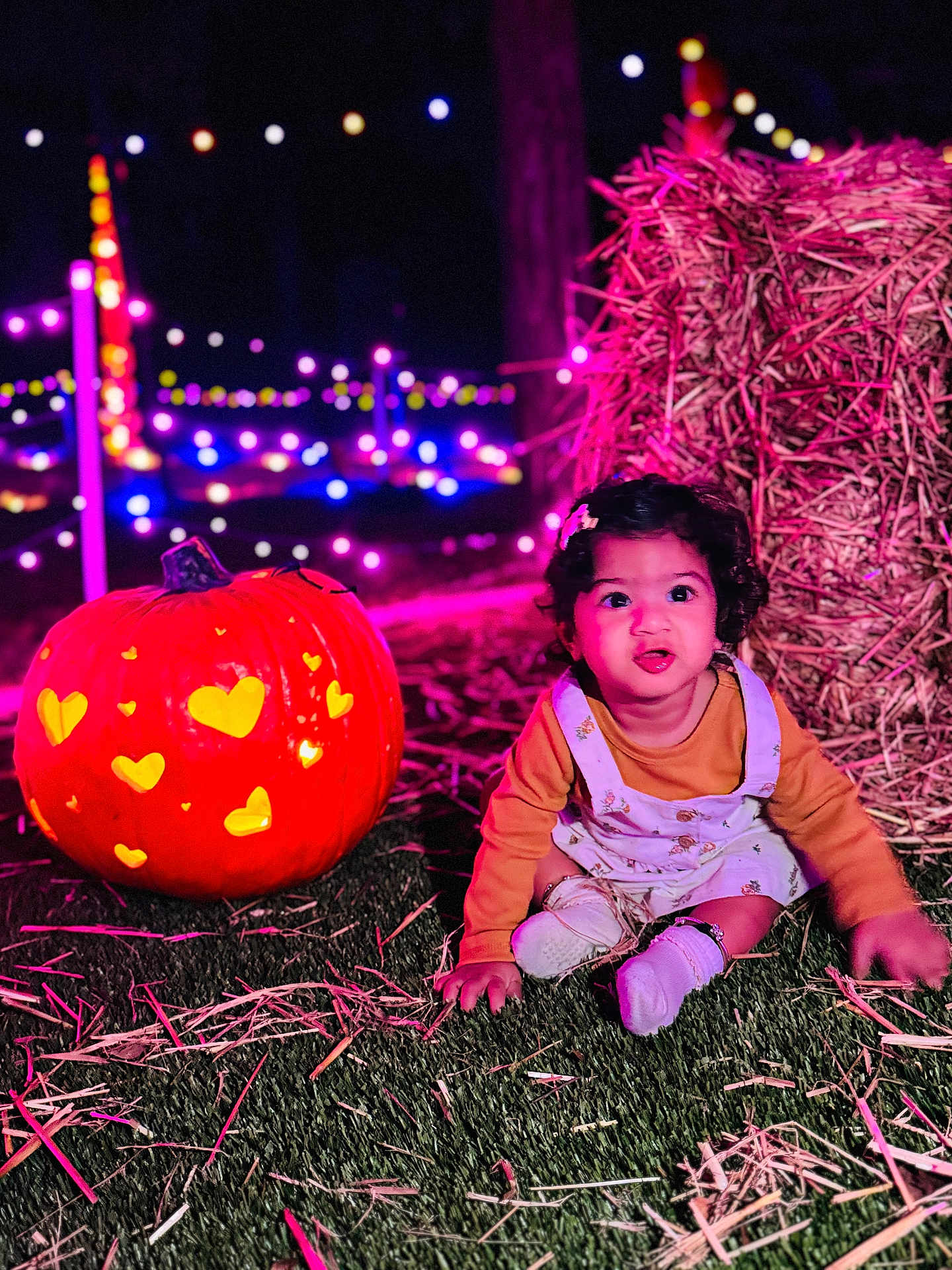 Tashvi is registered to the contest to win money with this photo: toddler, child, pumpkin, carved_pumpkin, grass, hay, night, string_lights, colorful_lights, festival, outdoor, cute, smiling, sitting, clothing, face, head, celebration, autumn, festive