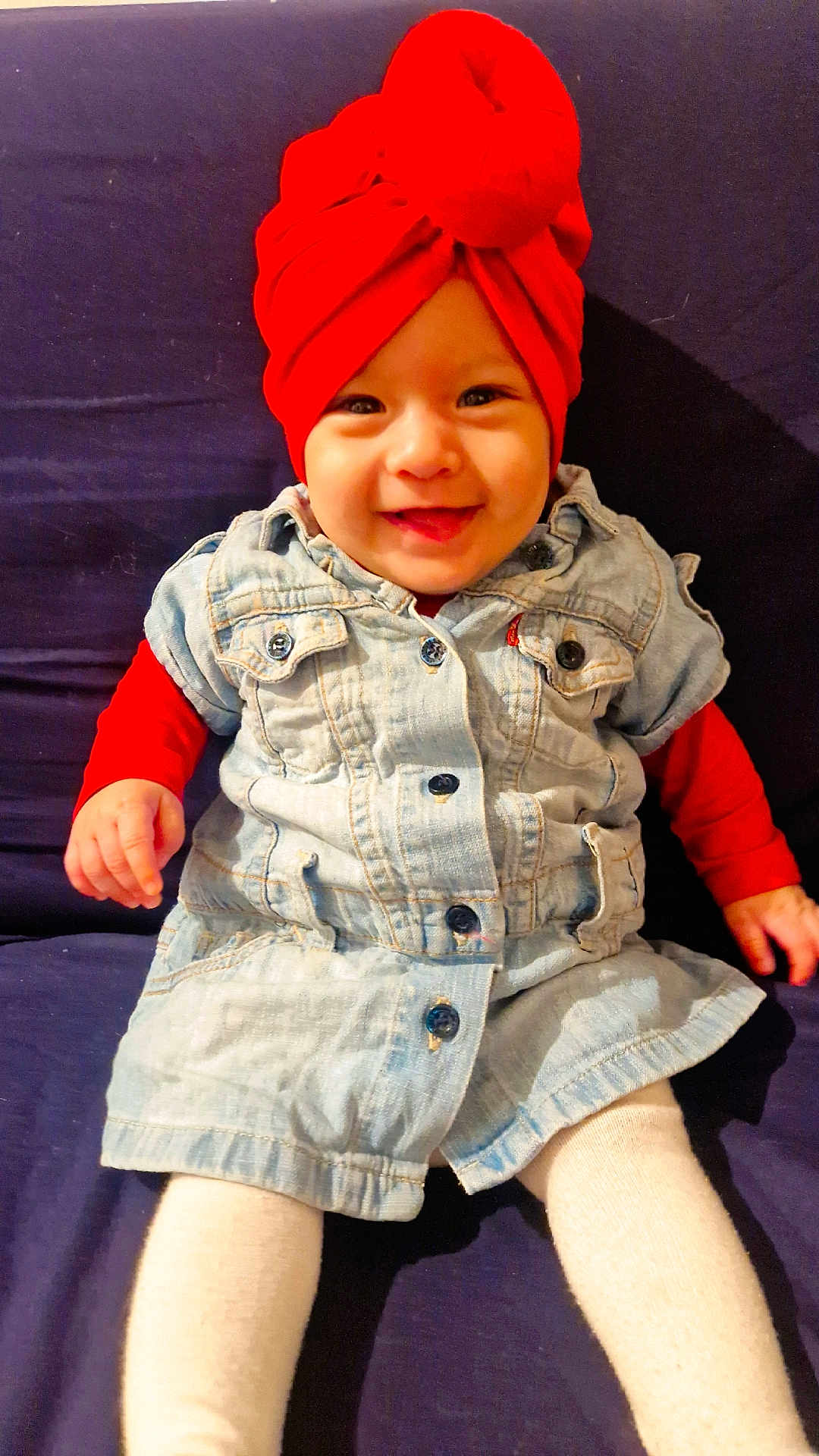 Ayline participe au concours pour gagner de l'argent avec cette photo : baby, infant, child, red_turban, headwrap, denim_dress, smiling, sitting, sofa, navy_background, white_tights, hands, eyes, portrait, cute, indoor, clothing, button, happy, fashion
