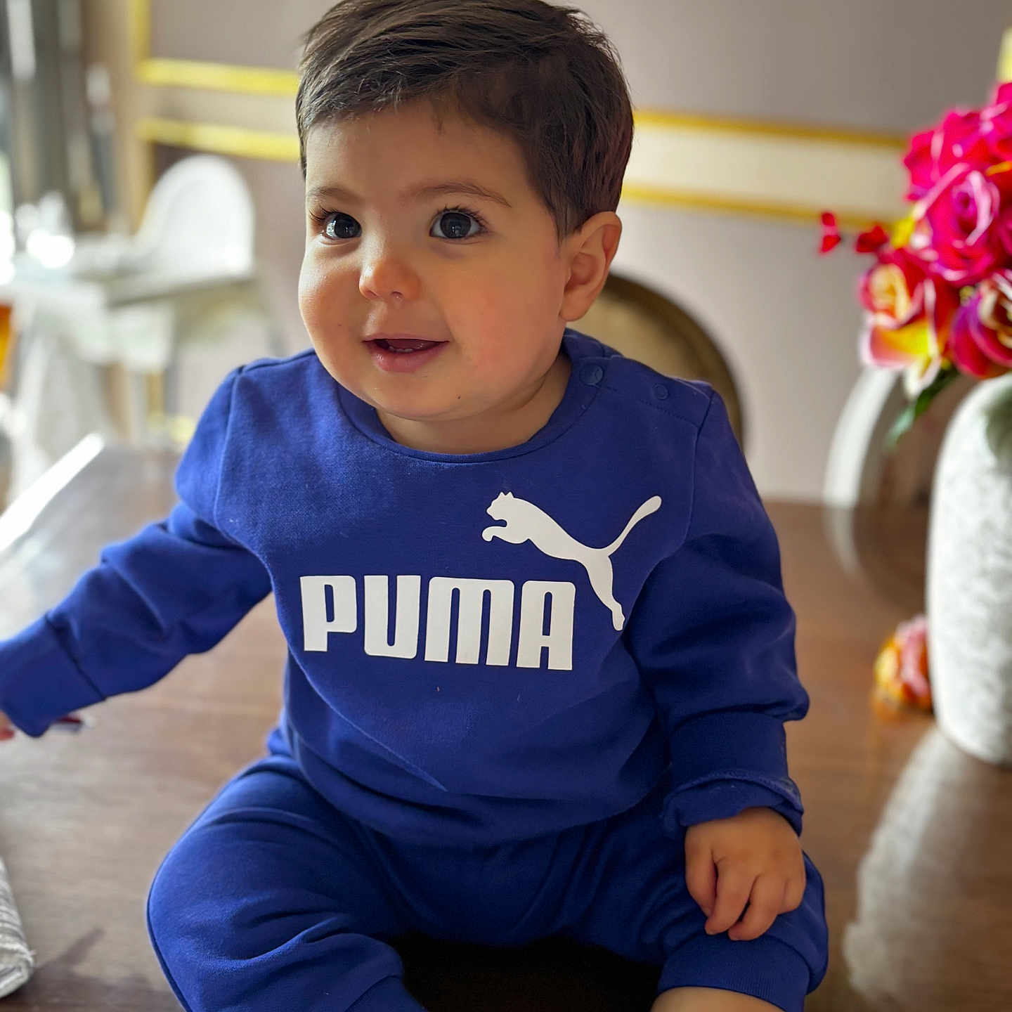Adam participe au concours pour gagner de l'argent avec cette photo : toddler, child, baby, blue_clothing, puma_logo, sitting, table, indoors, flowers, vase, curly_hair, smiling, cute, portrait, face, hand, socks, wooden_surface, decor, bright_colors