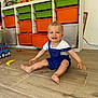 toddler, child, smiling, sitting, playroom, toys, storage_bins, colorful, wooden_floor, overalls, white_shirt, happy, indoor, face, blue_eyes, plastic_toys, whisk, floor, person, young_child