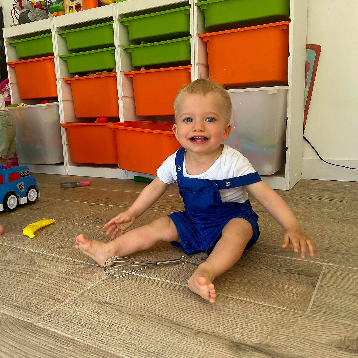 Eden participe au concours pour gagner de l'argent avec cette photo : blue_eyes, child, colorful, face, floor, happy, indoor, overalls, person, plastic_toys, playroom, sitting, smiling, storage_bins, toddler, toys, whisk, white_shirt, wooden_floor, young_child