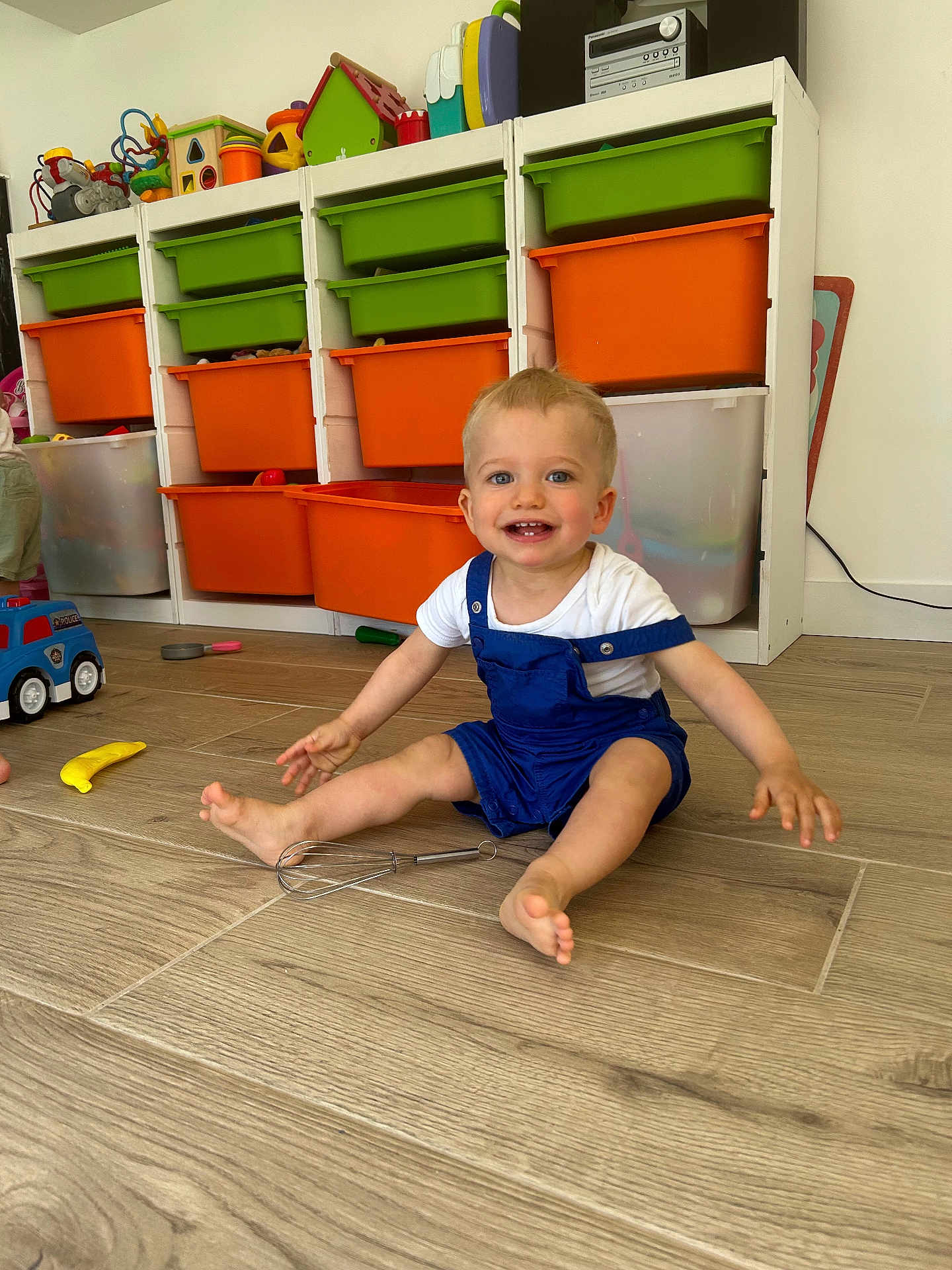 Eden participe au concours pour gagner de l'argent avec cette photo : toddler, child, smiling, sitting, playroom, toys, storage_bins, colorful, wooden_floor, overalls, white_shirt, happy, indoor, face, blue_eyes, plastic_toys, whisk, floor, person, young_child
