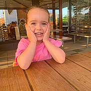 Ambre participe au concours pour gagner de l'argent avec cette photo : casual, child, daylight, face, girl, glass, hands_on_face, happy, indoor, joyful, natural_light, pink_shirt, portrait, restaurant, seated, smiling, table, window, wooden_table, young_child
