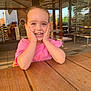 casual, child, daylight, face, girl, glass, hands_on_face, happy, indoor, joyful, natural_light, pink_shirt, portrait, restaurant, seated, smiling, table, window, wooden_table, young_child