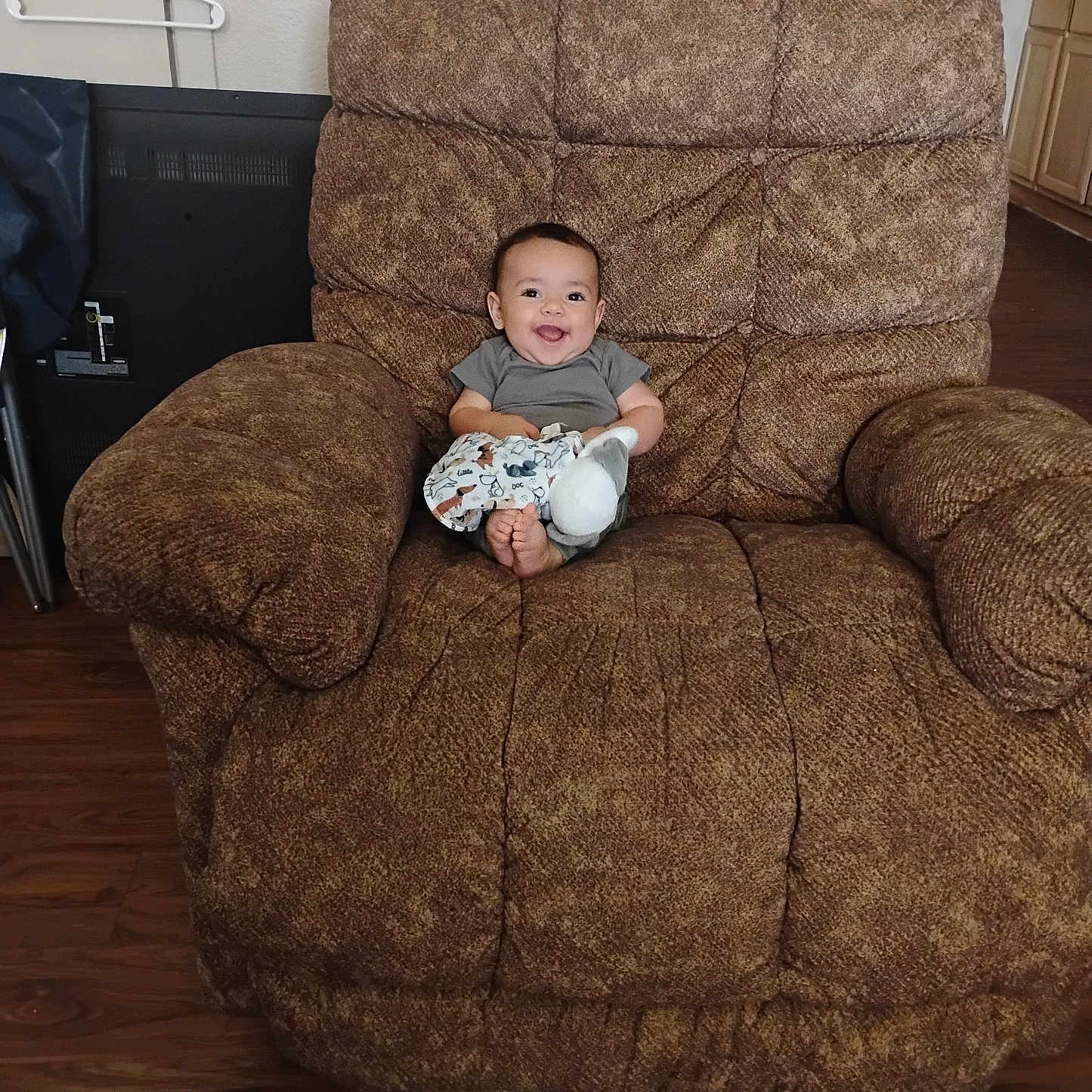 Corben joined the competition — help win amazing prizes! baby, brown_chair, child, clothing, cozy, cute, floor, furniture, hanger, happy, home, indoor, infant, person, recliner, seated, smiling, stuffed_toy, wall, wood_floor